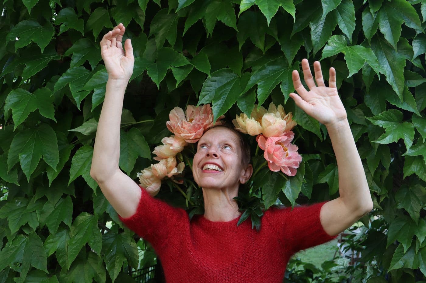 Heide hebt lachend beide Arme vor einer Wand aus wildem Wein